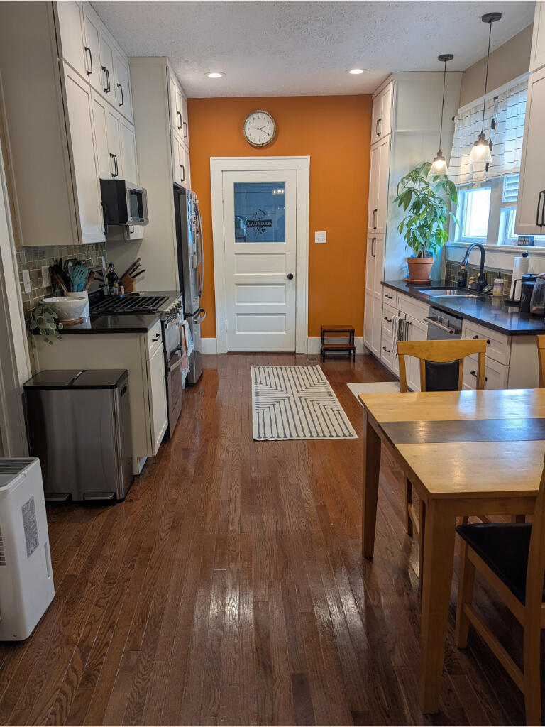 A warm, lived-in kitchen with orange walls, brown wood floors, and soft lighting. The space includes a clean island, seasonal decor, and a welcoming dining area in the background. It feels personal, vibrant, and freshly maintained.